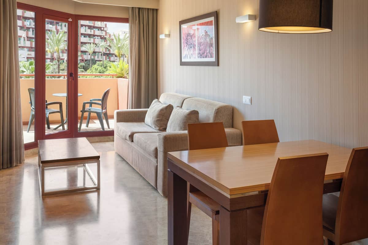 Een woonkamer met een bank, een eettafel en stoelen, een balkon met uitzicht op gebouwen en palmbomen, en een lijst met een foto aan de muur.