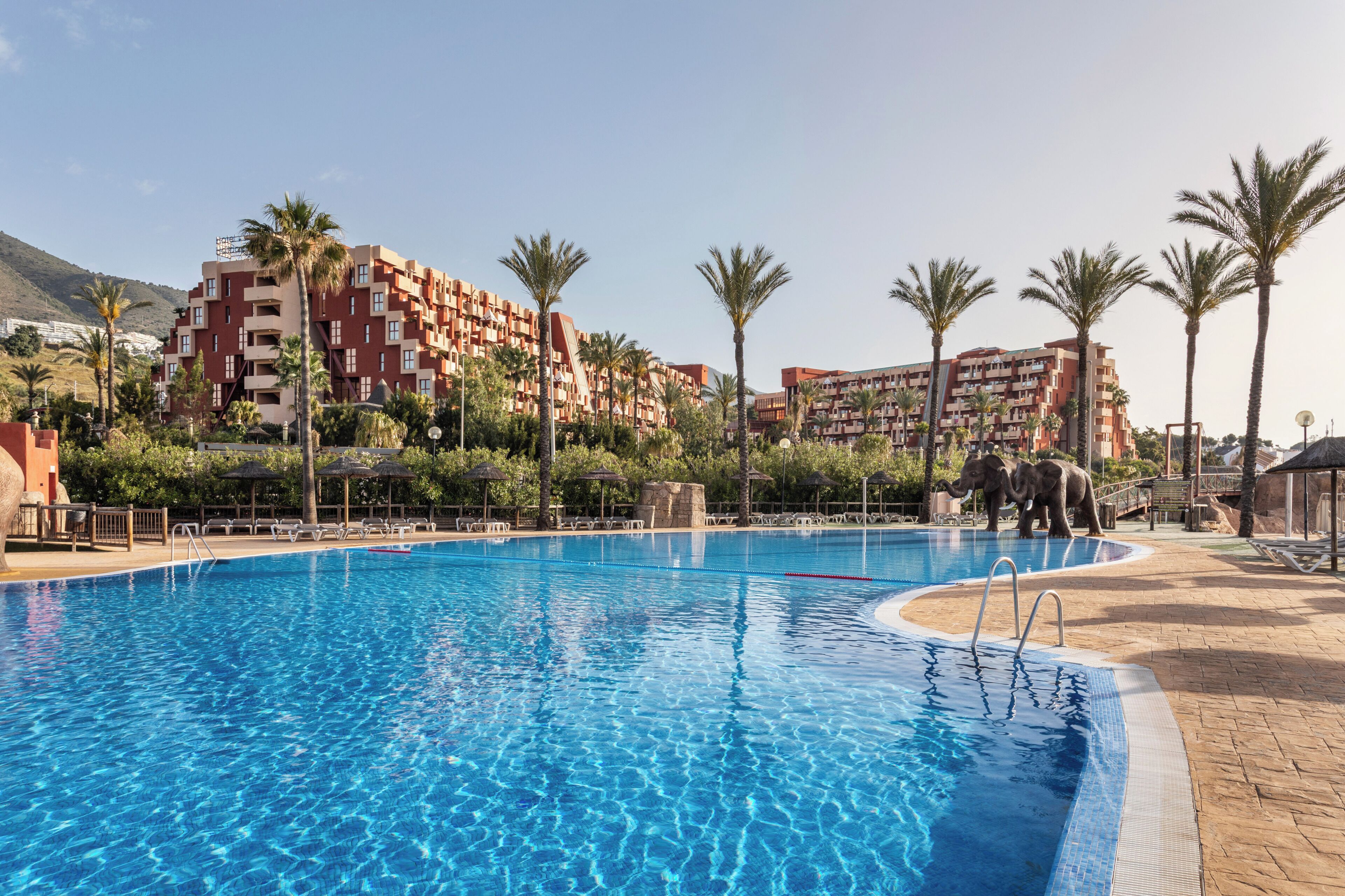A swimming pool with a clear blue water, surrounded by palm trees and a building with balconies.