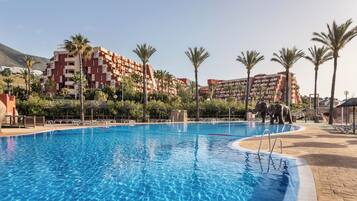 A swimming pool with a clear blue water, surrounded by palm trees and a building with balconies.