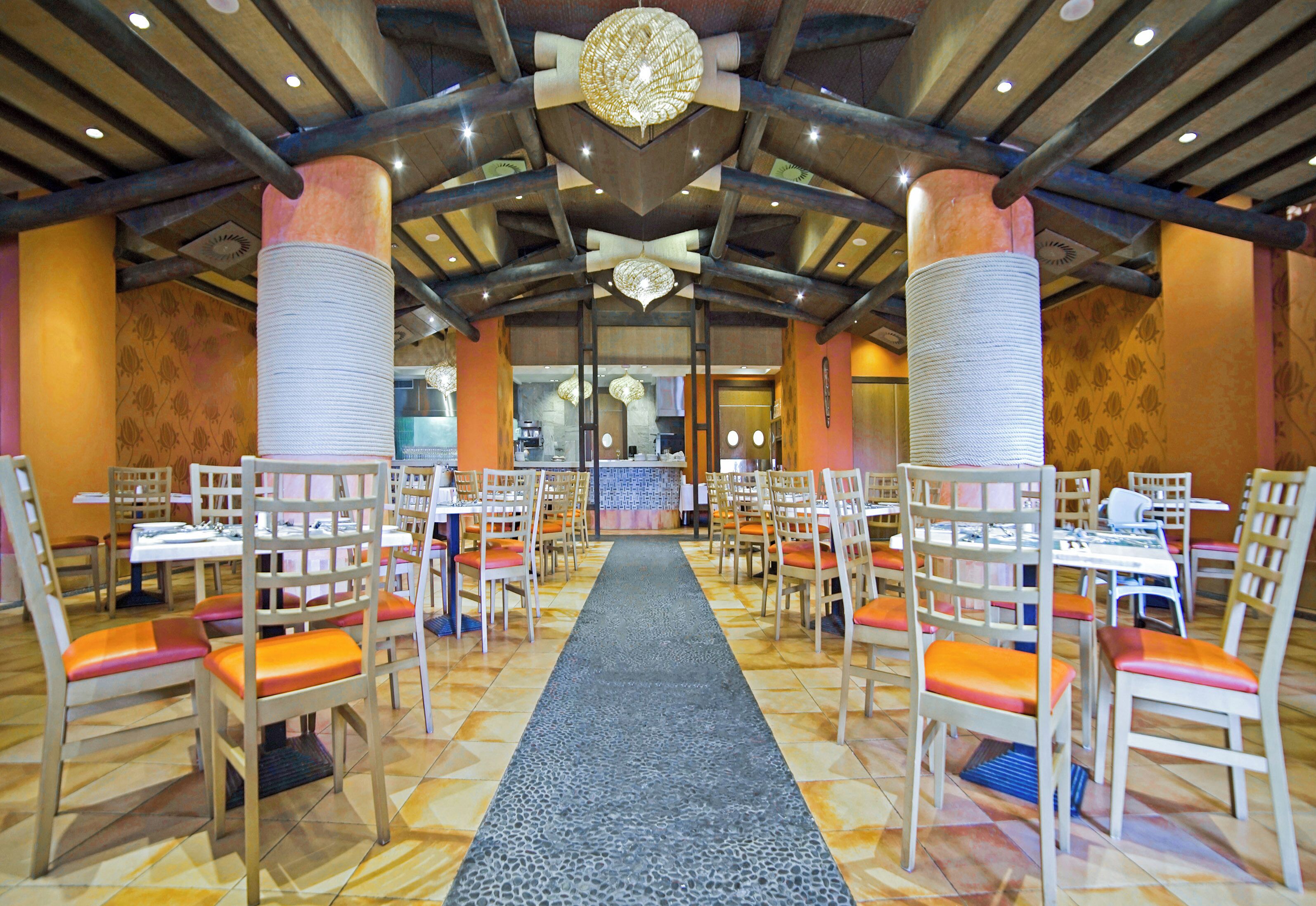 A restaurant with a central aisle, wooden chairs with orange cushions, and a patterned carpet.