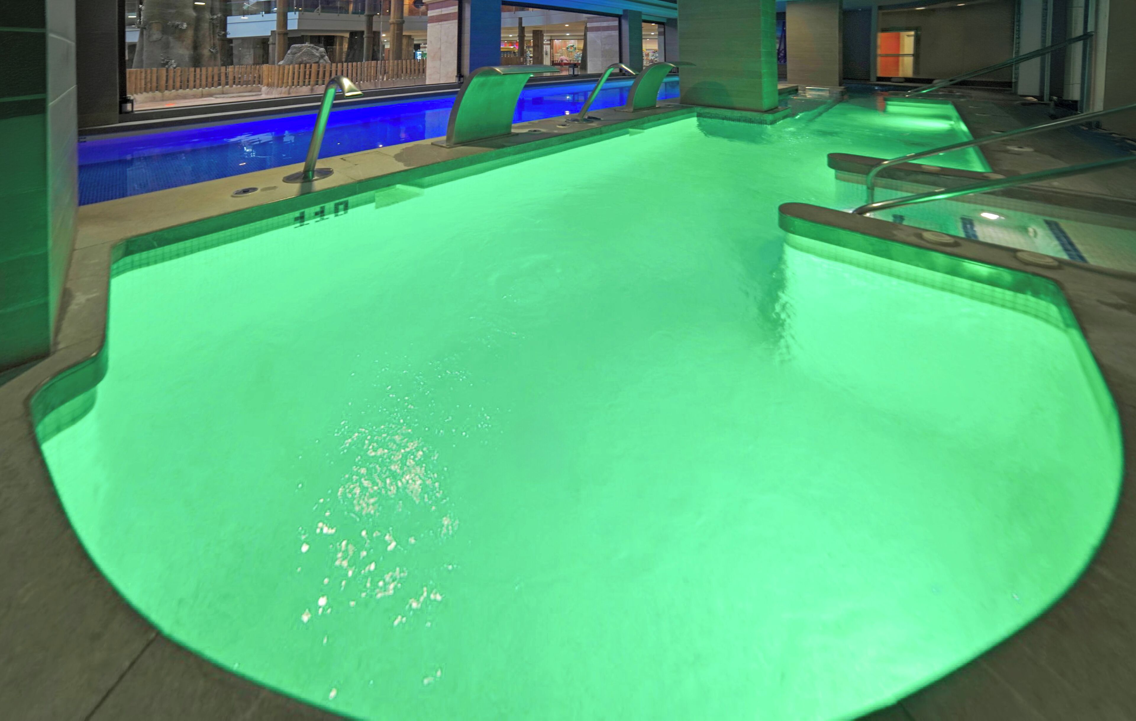 A large indoor swimming pool with a greenish hue, surrounded by a concrete deck and metal railings.