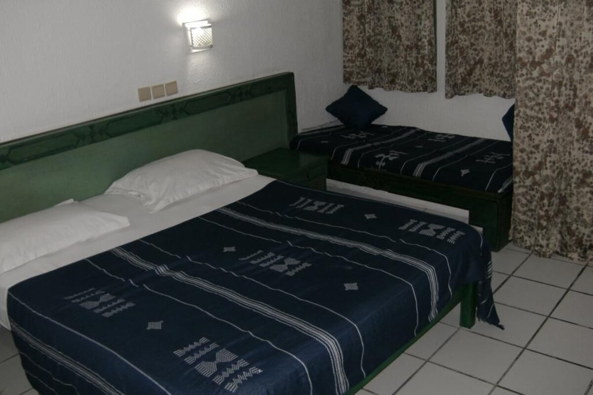 A room with two beds, a green headboard, a wall-mounted light, and a patterned curtain.