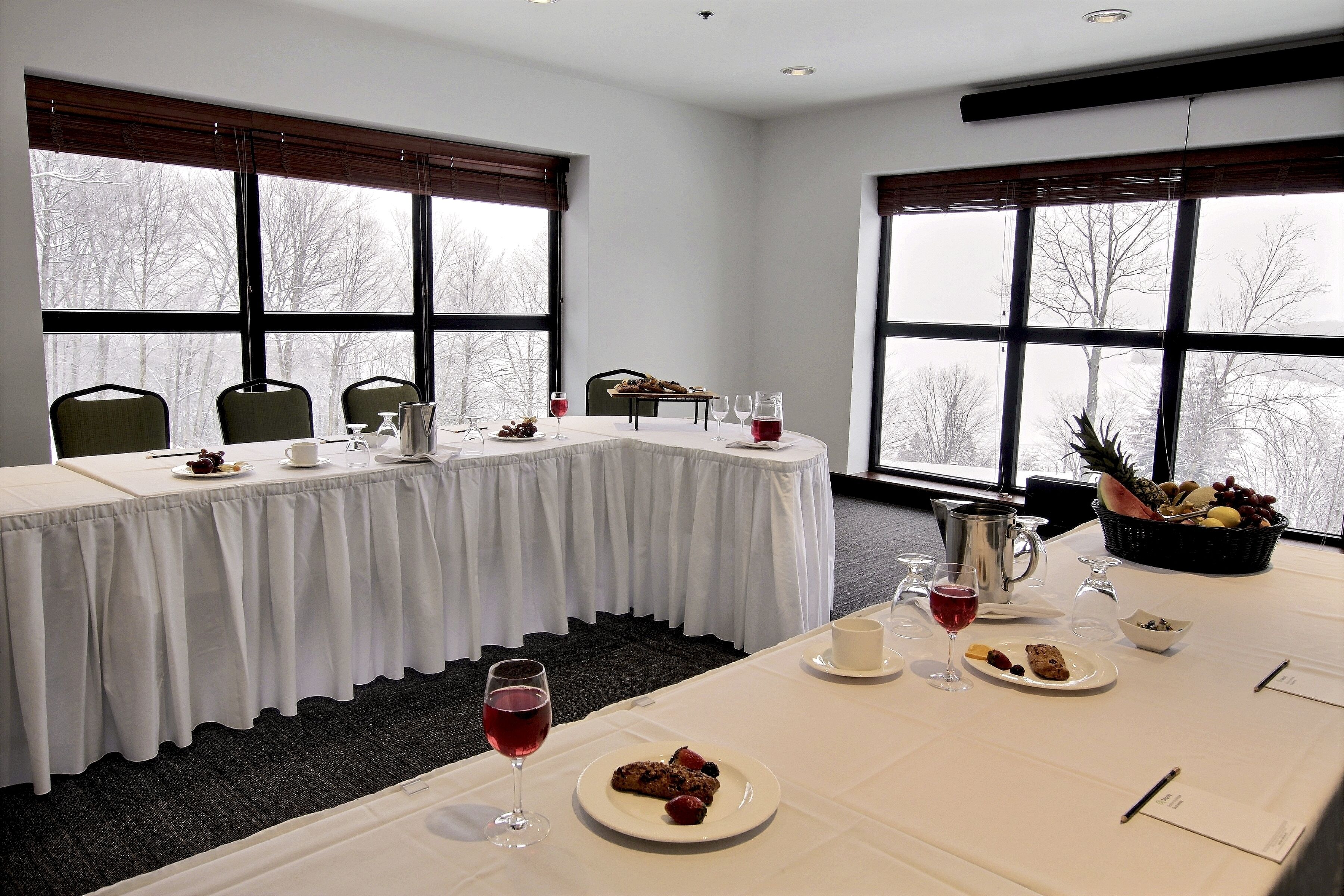 Un salón de conferencias con una larga mesa dispuesta para una reunión, ventanas amplias con vistas a árboles y una cesta de frutas sobre la mesa.