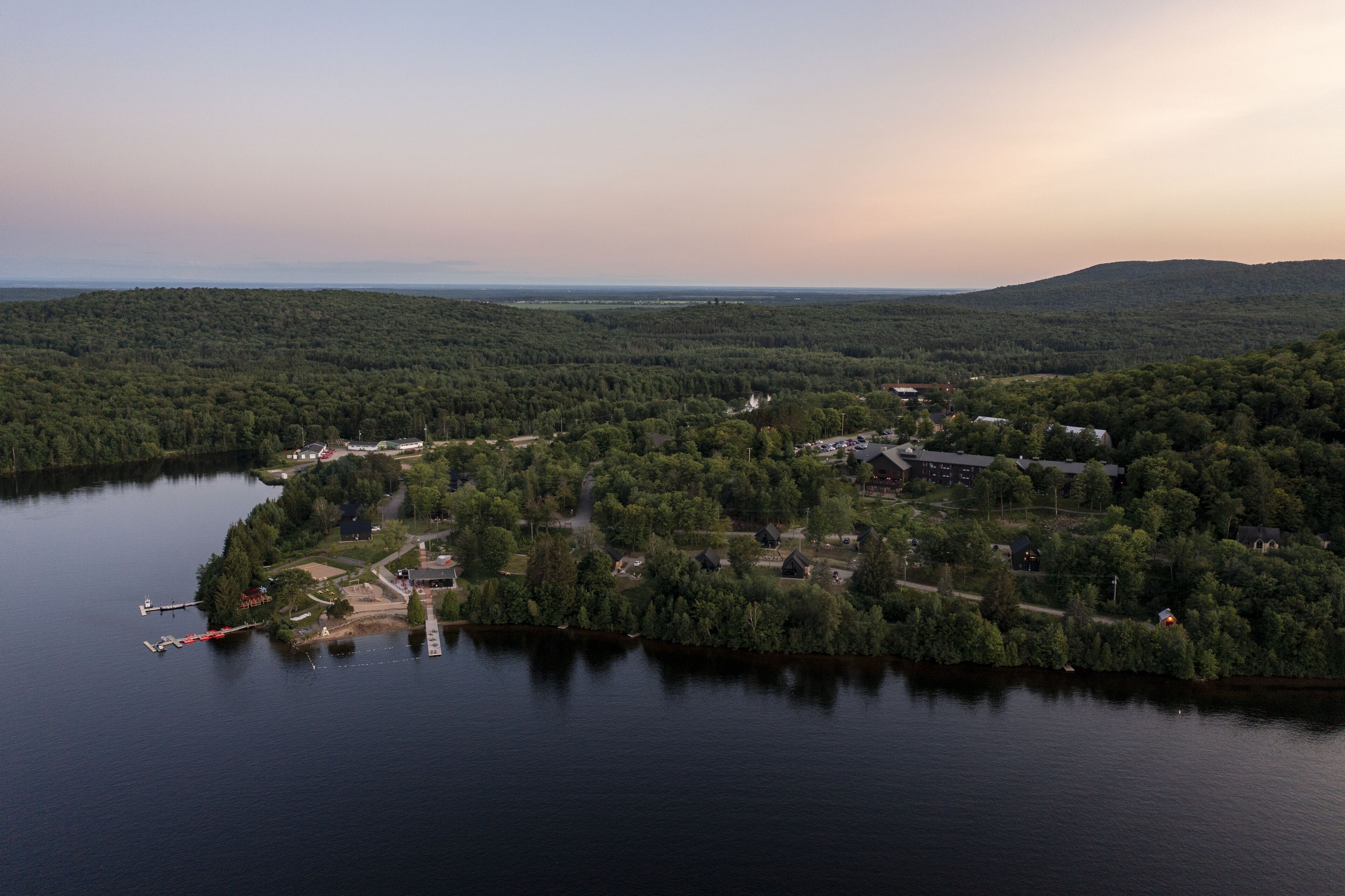 An aerial view of a lakeside property with a dock, surrounded by dense forest and hills in the background.