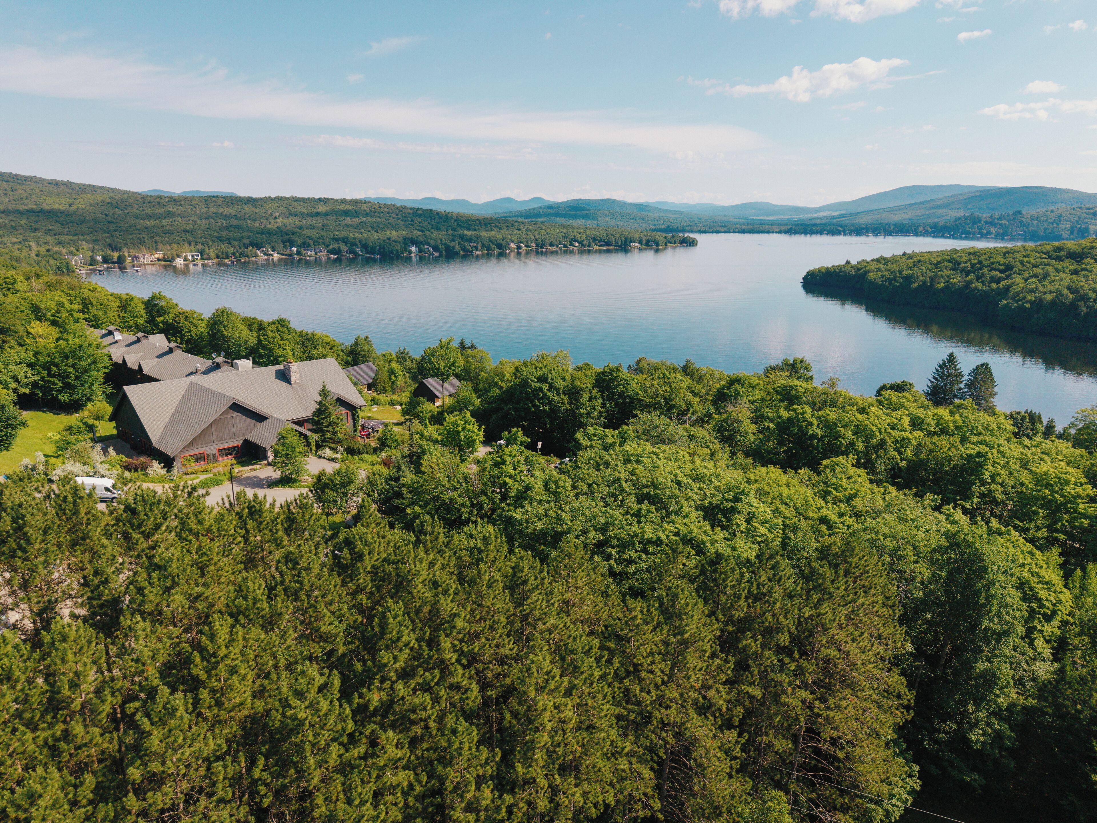A lakeside property with multiple buildings, surrounded by dense forest and mountains in the background.