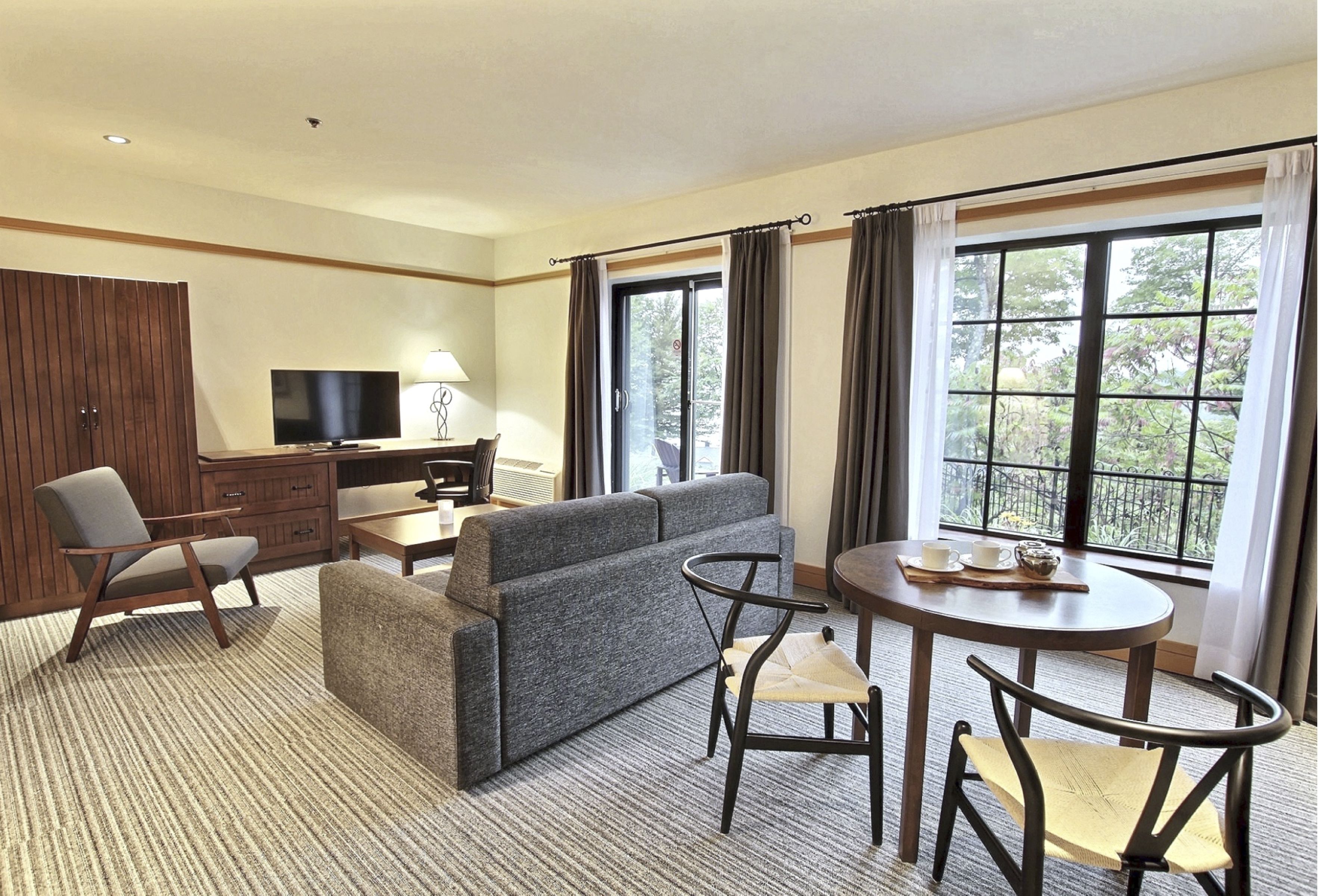 Una habitación de hotel moderna con sofá, sillas, televisor, mesa de comedor y ventanas amplias con cortinas.