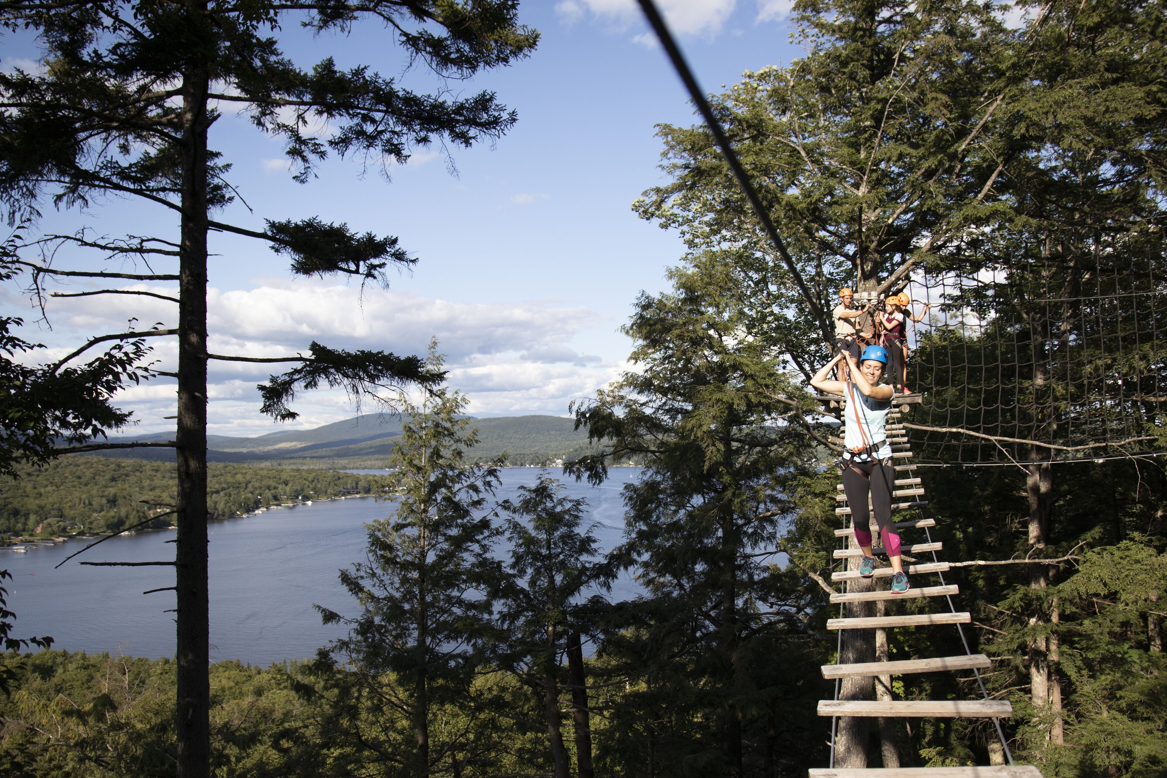 A person navigating a rope bridge in a forested area with a lake and mountains in the background.