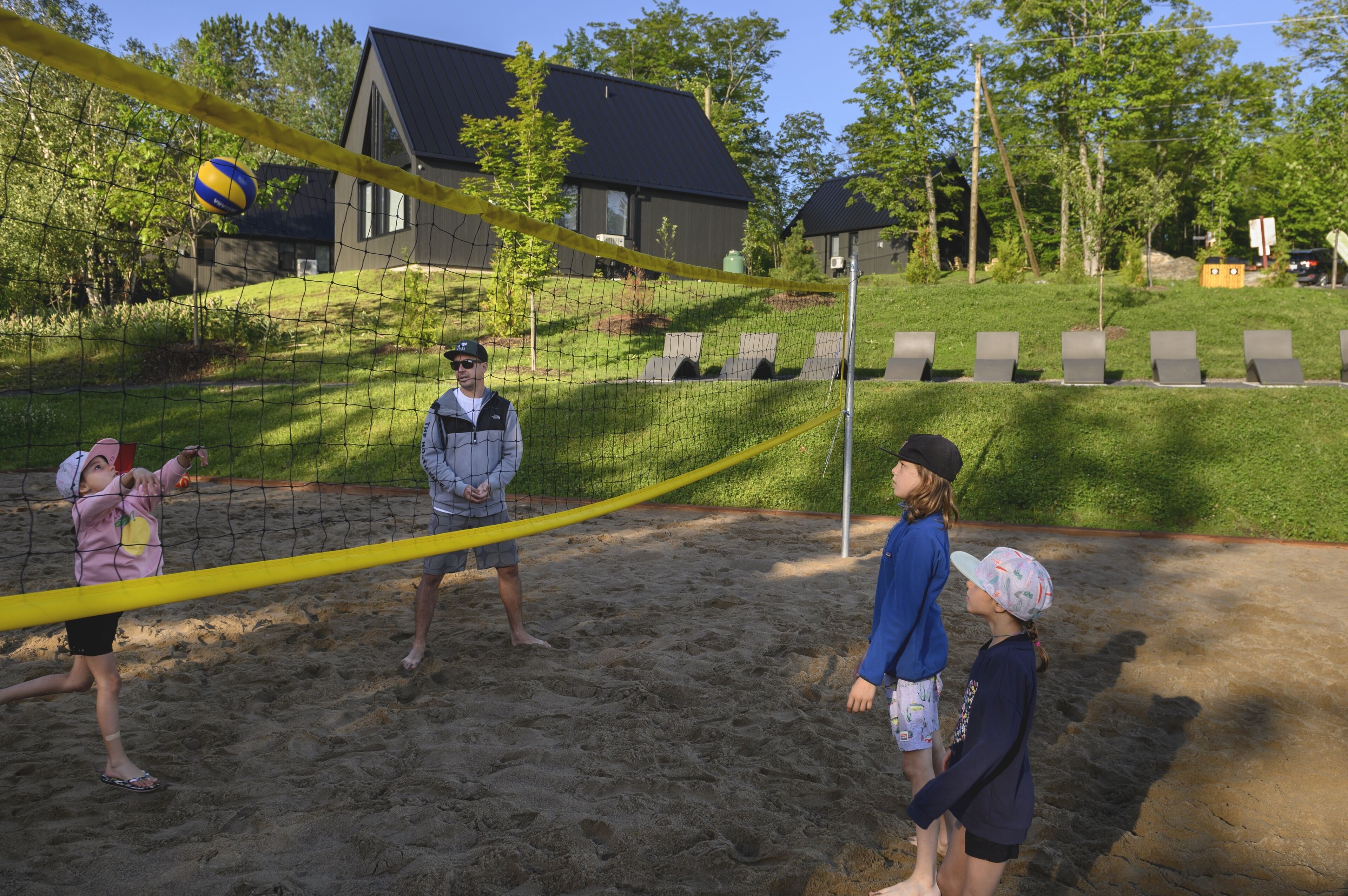 A volleyball game on a sandy court with a net, surrounded by a grassy area and a building in the background.