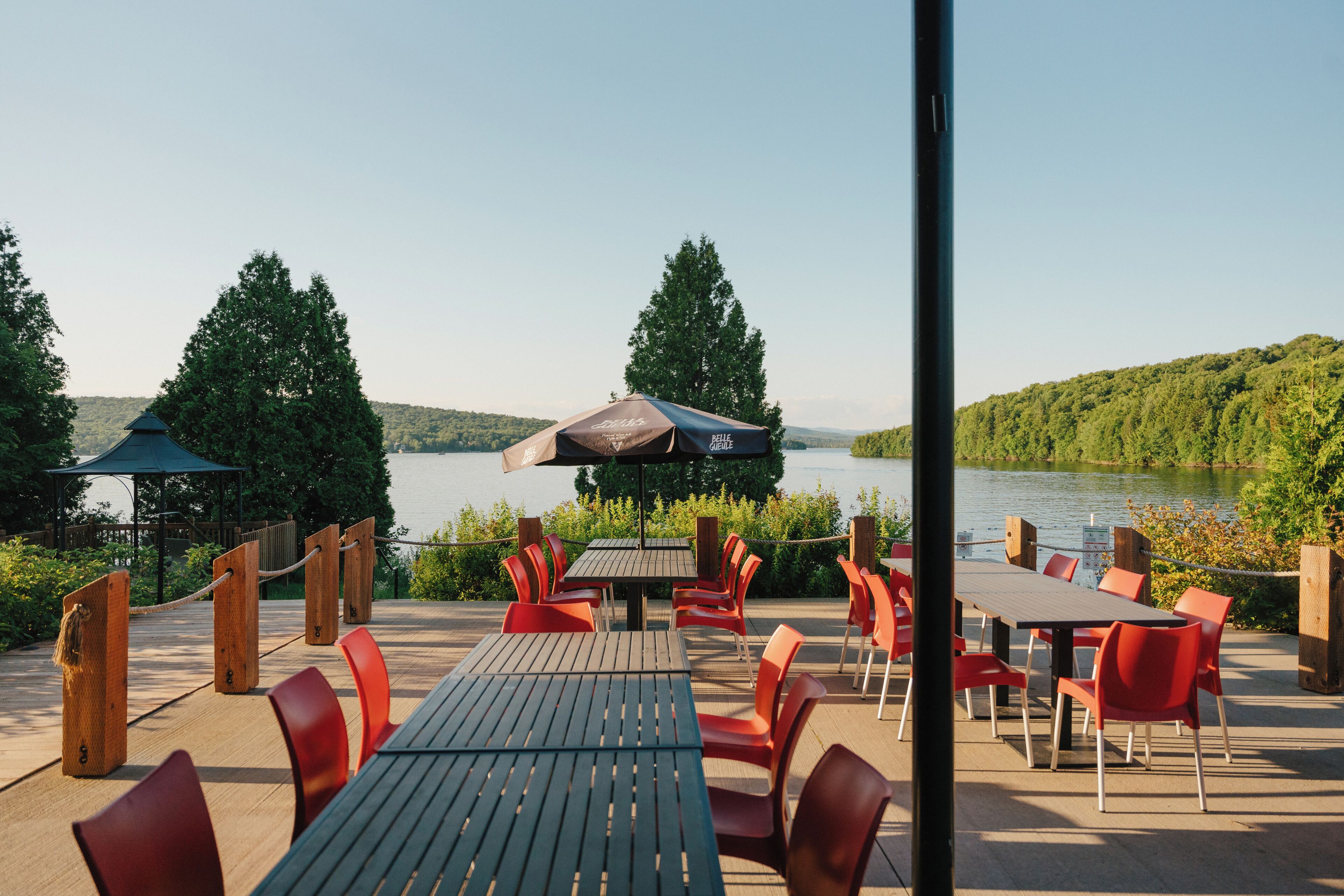 An outdoor dining area with tables and chairs, overlooking a body of water and surrounded by greenery.