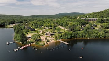 Vue aérienne d’un complexe hôtelier au bord d’un lac, avec un quai, des bateaux et un bâtiment entouré de collines boisées.