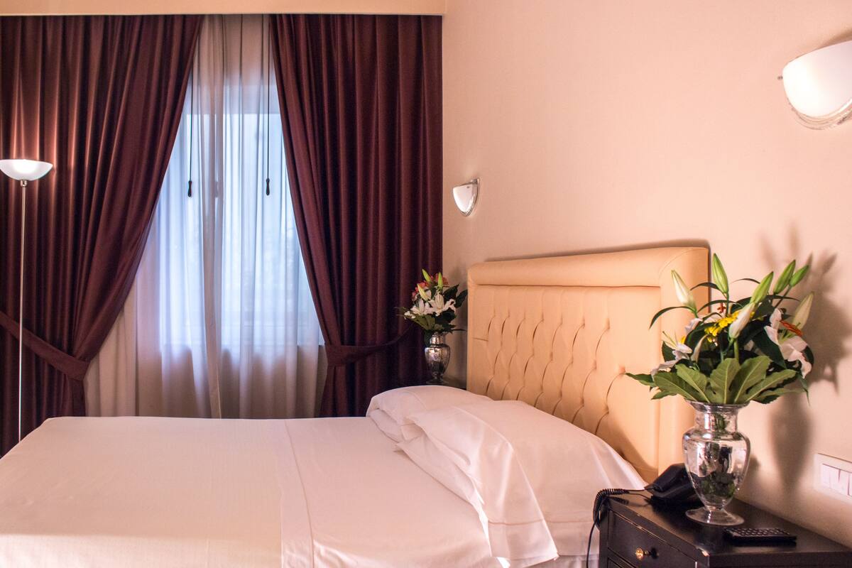Une chambre d’hôtel avec un lit, une table de chevet, une lampe, un vase avec des fleurs et des rideaux.