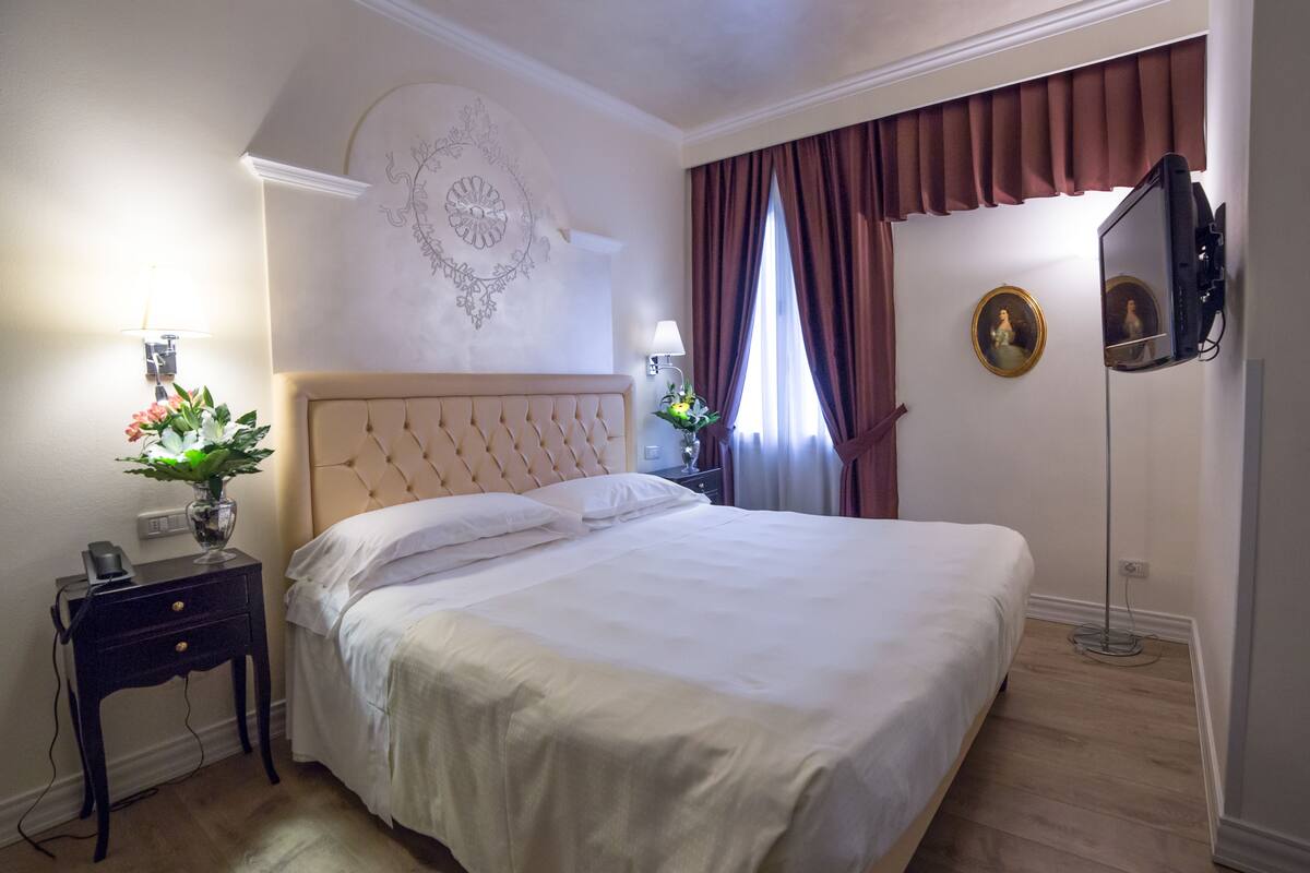 Un lit bien fait, avec une tête de lit, une table de chevet avec une lampe et un téléviseur fixé au mur.