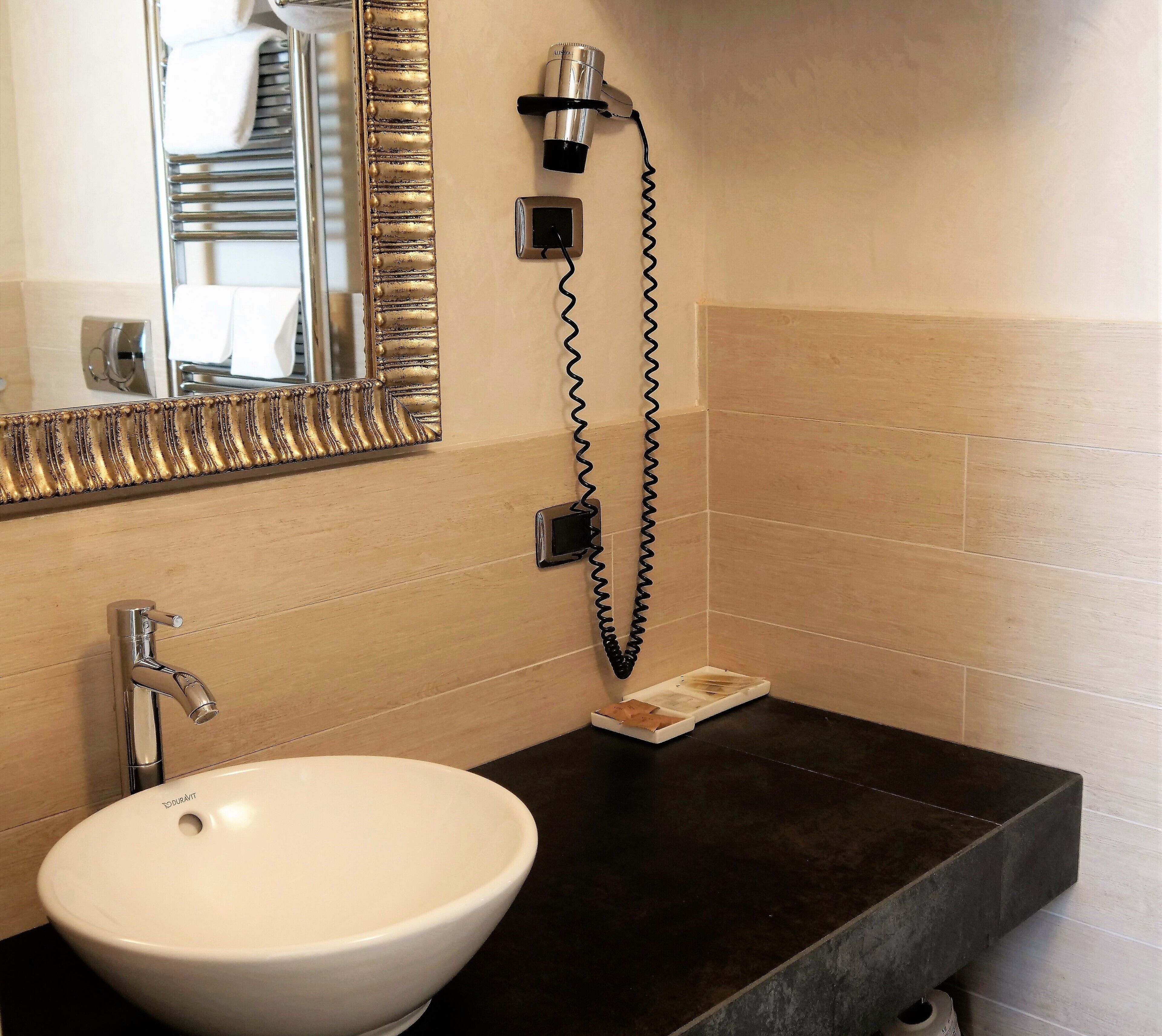 Une salle de bain avec un lavabo blanc, un miroir avec un cadre doré, un sèche-cheveux et une petite poubelle.