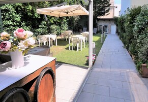 Un patio al aire libre con mesas y sillas, una mesa de madera con un jarrón de flores, y un camino que lleva a un edificio.