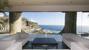 A modern interior with a large window offering a coastal view, including a beach and buildings on a cliff. - Gloria Palace Royal Hotel & Spa (Mogan)