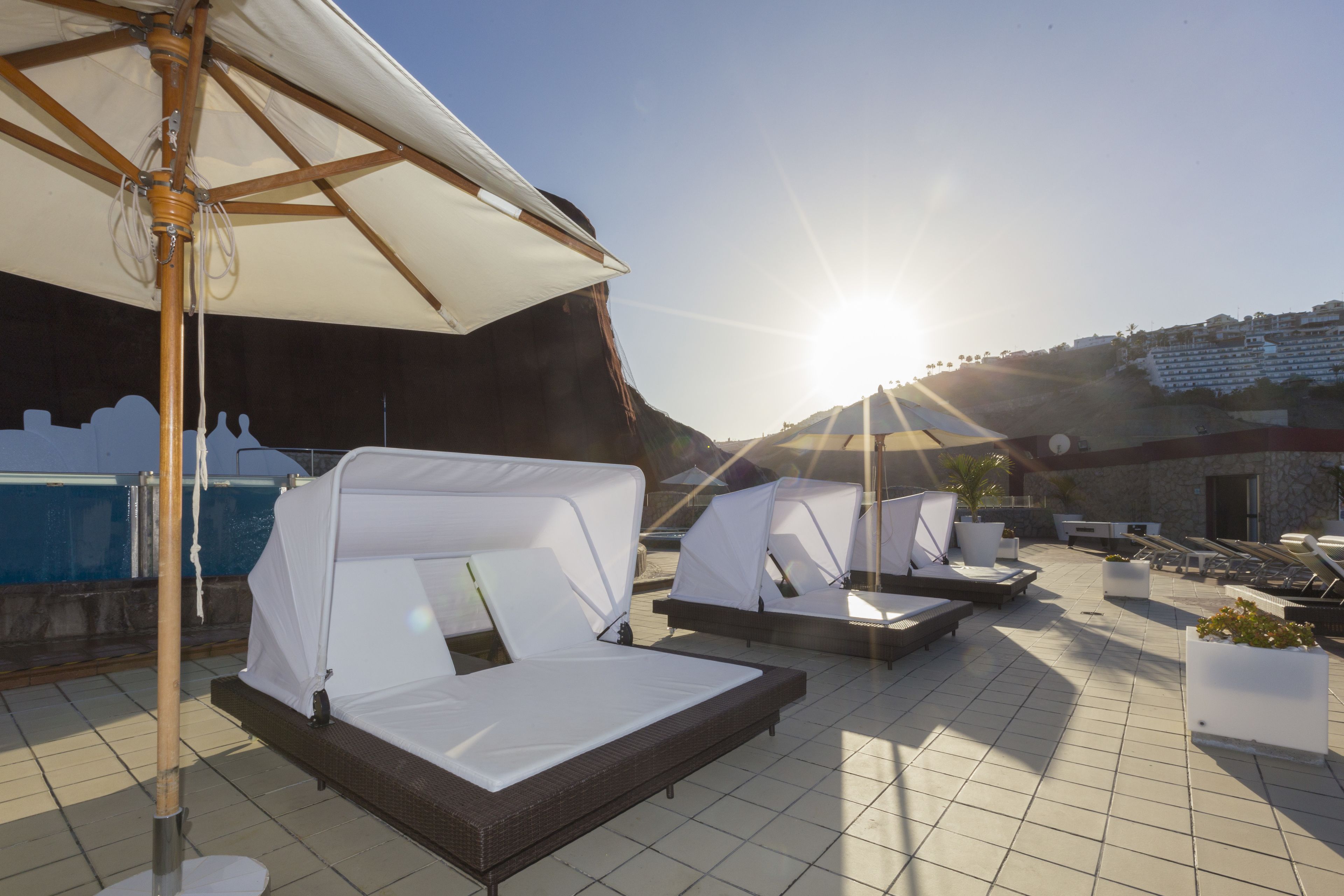A rooftop lounge area with sunbeds and umbrellas, overlooking a pool and a building in the background.