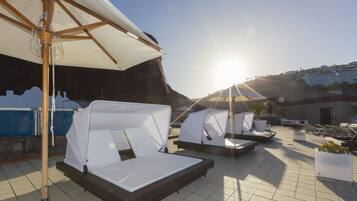 A rooftop lounge area with sunbeds and umbrellas, overlooking a pool and a building in the background.