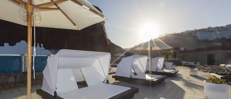 A rooftop lounge area with sunbeds and umbrellas, overlooking a pool and a building in the background.