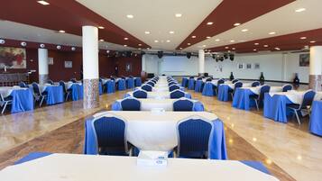 Un salón de conferencias con filas de mesas y sillas, paredes blancas y un escenario al frente.