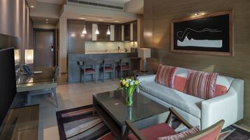 A modern hotel room with a living area, a glass coffee table, a sofa with striped cushions, a TV, and a kitchen area with bar stools.