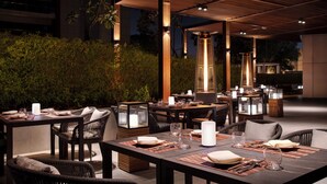 Een terras met tafels en stoelen, sfeerverlichting en een uitzicht op een gebouw.