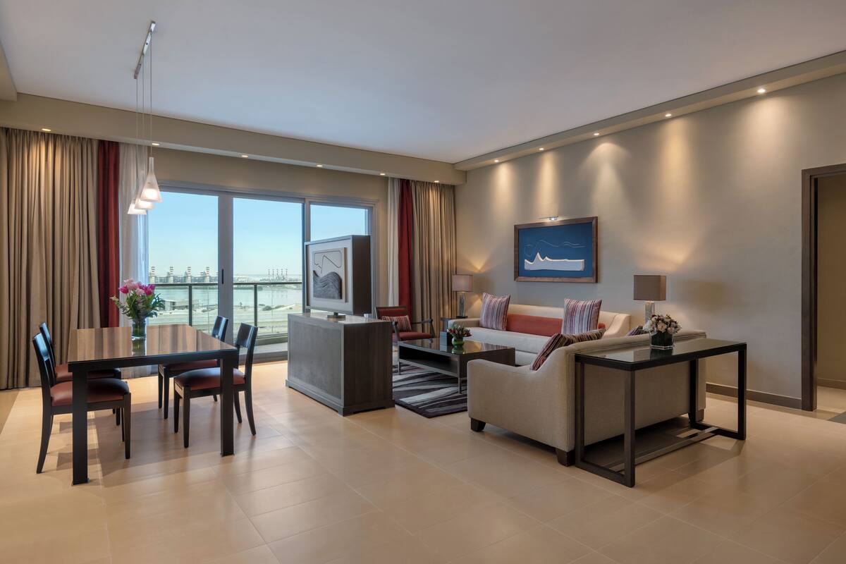 A modern hotel room with a dining area, a sofa, a TV, and a large window with a city view.