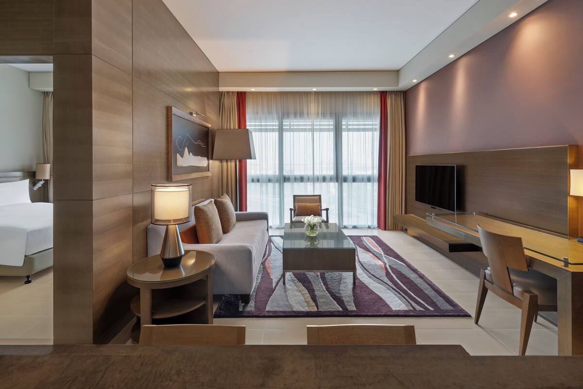 A modern hotel room with a bed, a sofa, a coffee table, a TV, and a desk area.