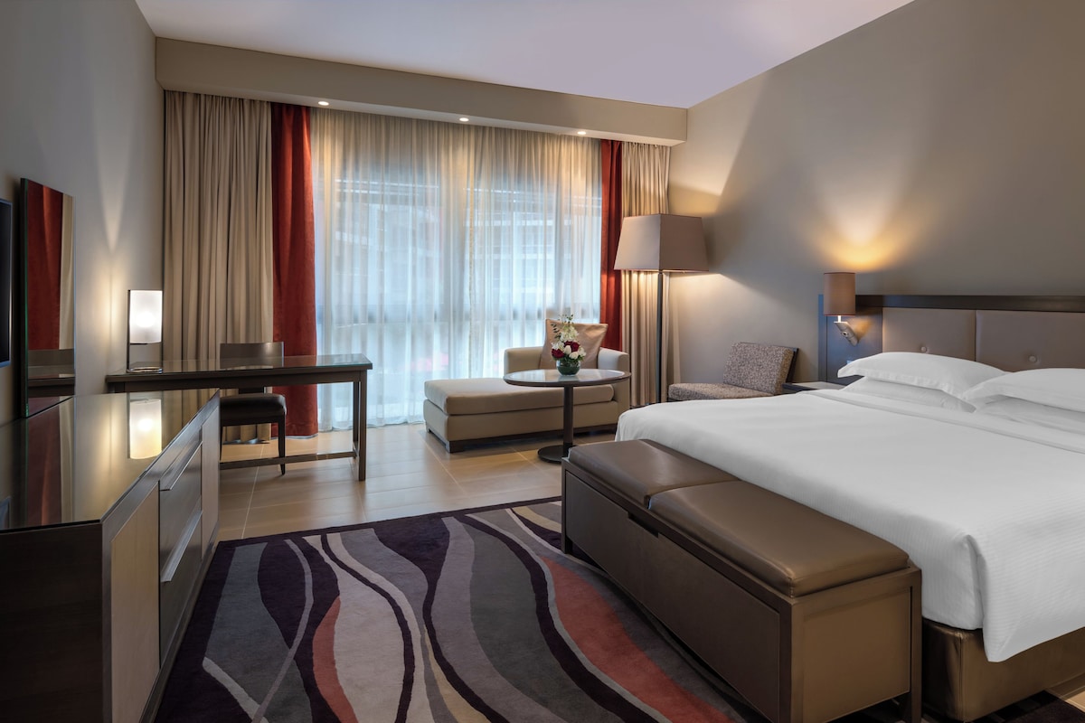 Une chambre d’hôtel avec un lit, un bureau, une chaise, une petite table avec un vase et une lampe.