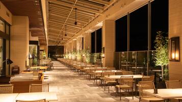 A spacious indoor dining area with multiple tables and chairs, large windows, and a high ceiling with exposed beams.