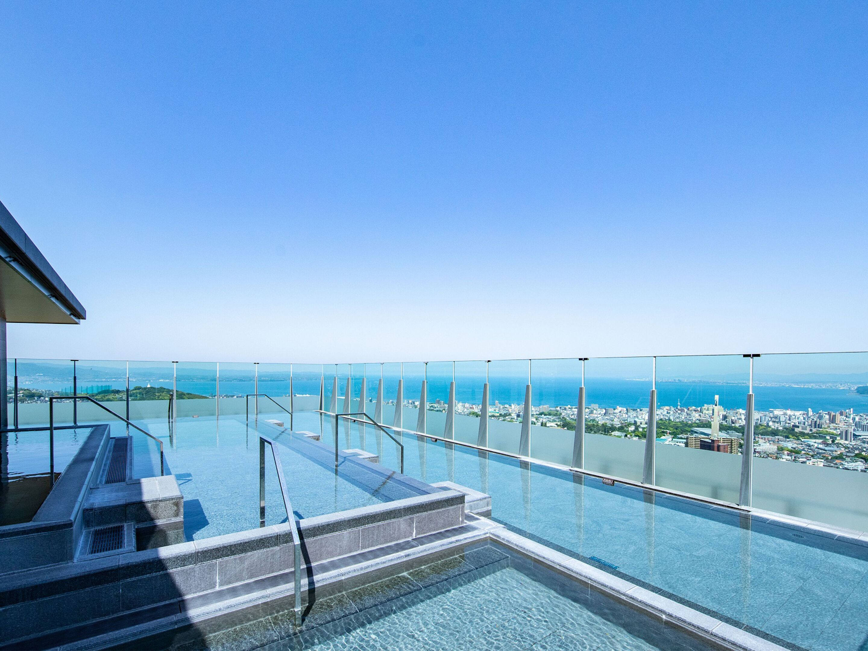 A rooftop infinity pool with glass railings overlooking a cityscape and the sea.