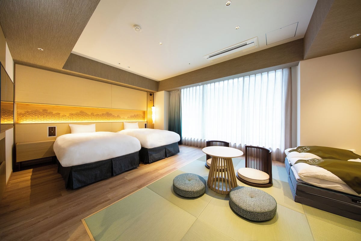 Hotellihuone, jossa on kaksi sänkyä, oleskelualue pöydällä ja tuoleilla sekä suuri ikkuna, jossa on verhot.