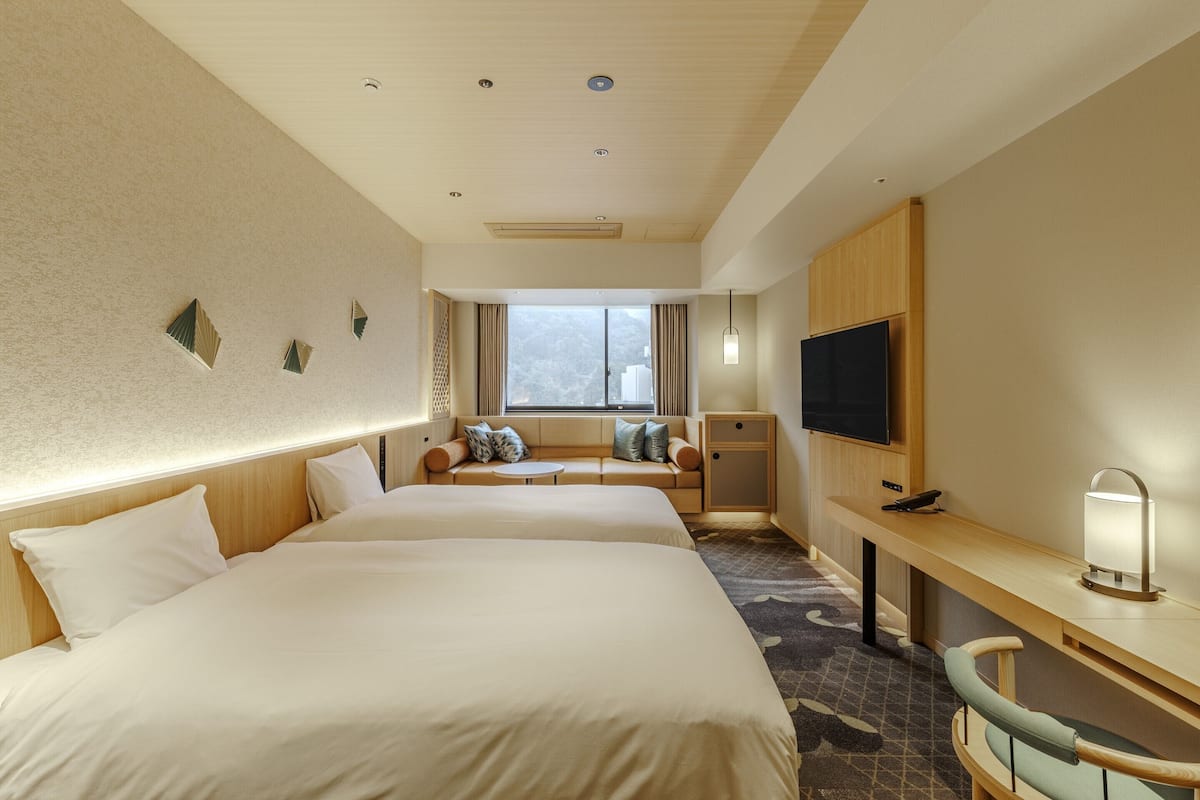 Hotellihuone, jossa on kaksi sänkyä, sohva, työpöytä, televisio ja ikkuna, josta on näkymä.