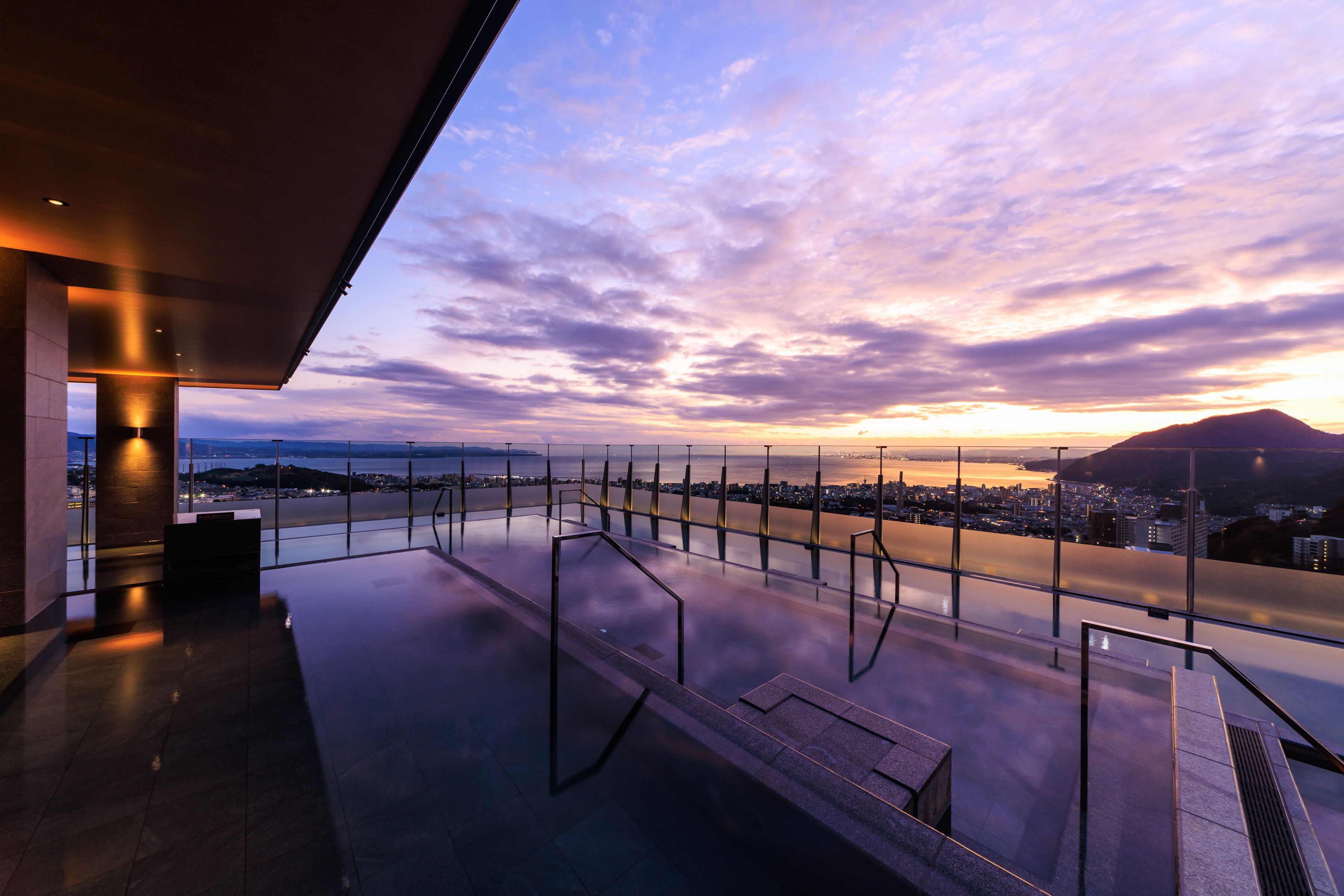 A modern building with a glass facade overlooking a cityscape and a body of water at sunset.