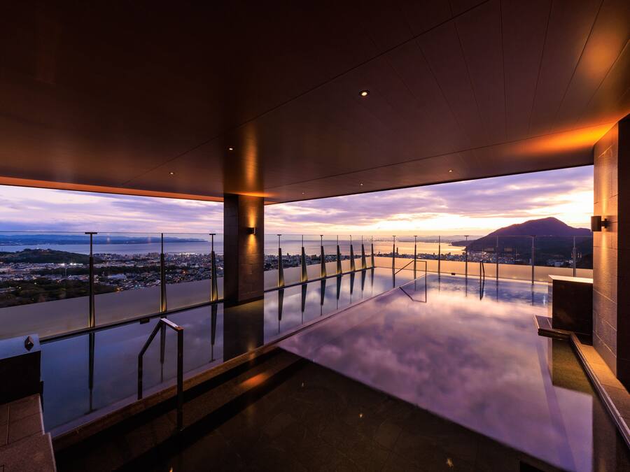 A modern hotel room with a large glass balcony overlooking a cityscape and a body of water.