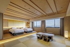 A hotel room with a bed, bedside tables, a seating area with a table and chairs, and a large window with a view.