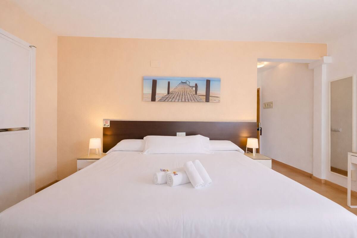 A hotel room with a large bed, two bedside tables with lamps, a painting on the wall, and a door leading to another room.