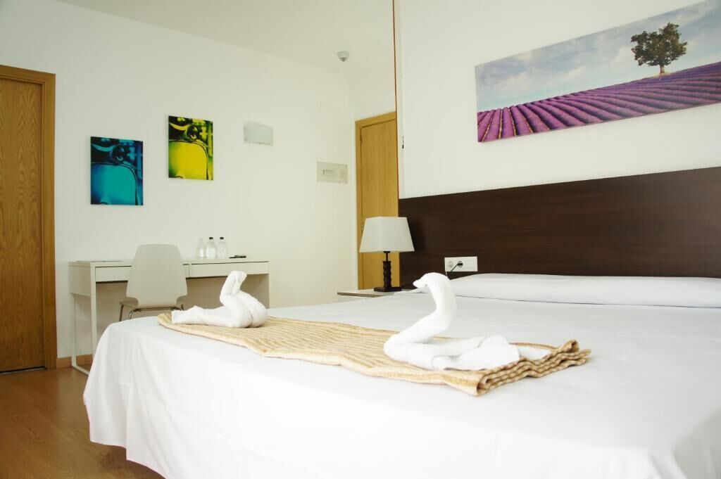 A hotel room with a bed, a desk, a chair, and a painting on the wall.