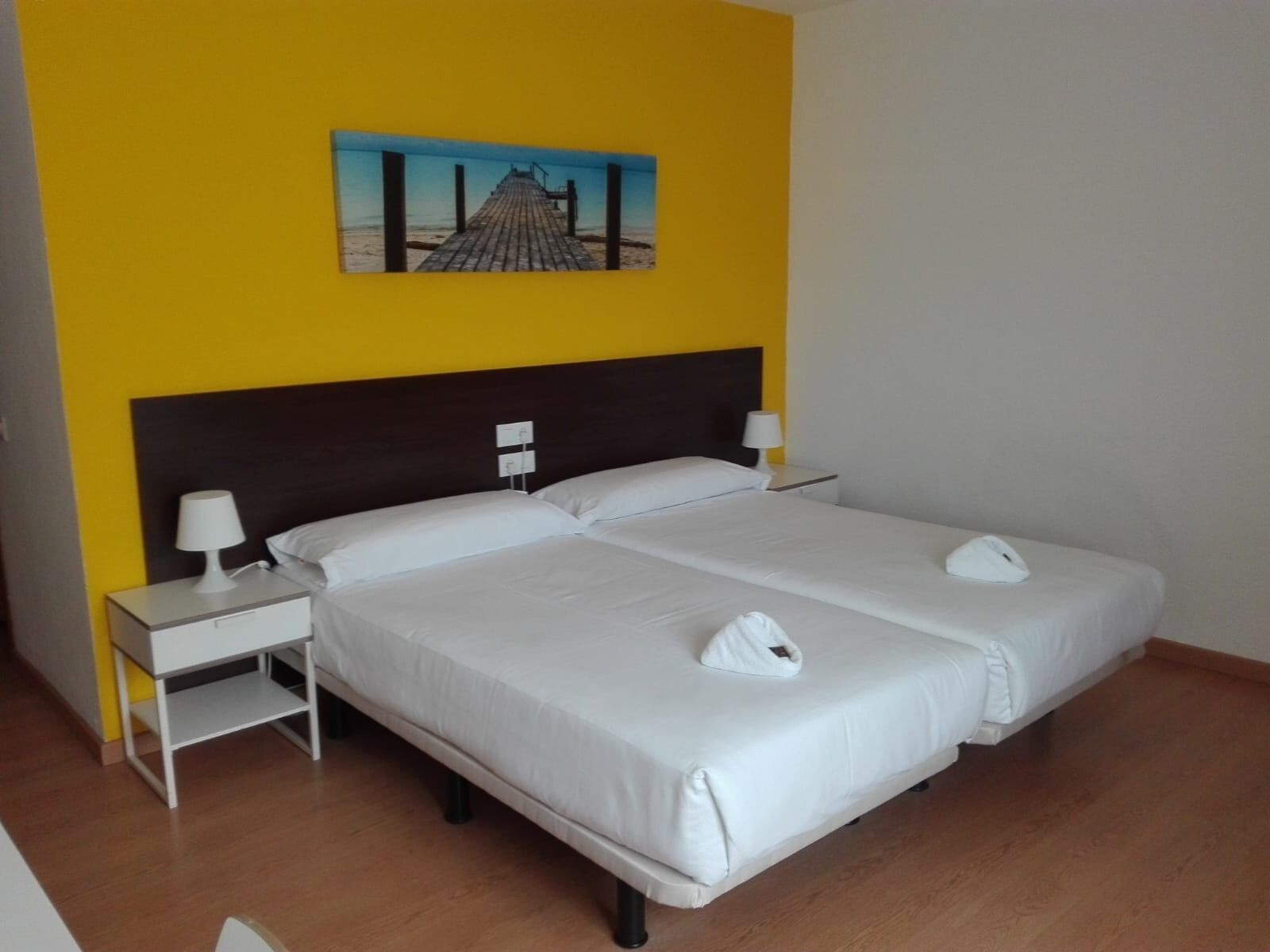 A hotel room with a double bed, two bedside tables with lamps, a headboard, and a painting of a pier.