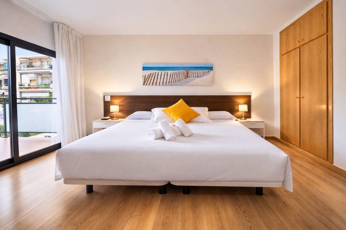 A modern hotel room with a large bed, wooden headboard, and a view of the city through floor-to-ceiling windows.