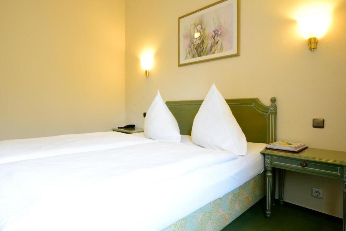 A hotel room with a bed, two bedside lamps, a nightstand with a book, and a wall-mounted painting.