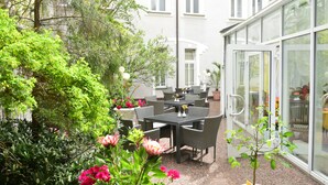 An outdoor patio with a table and chairs, surrounded by potted plants and a glass door leading to a building. - Hotel Alexandra (Plauen)
