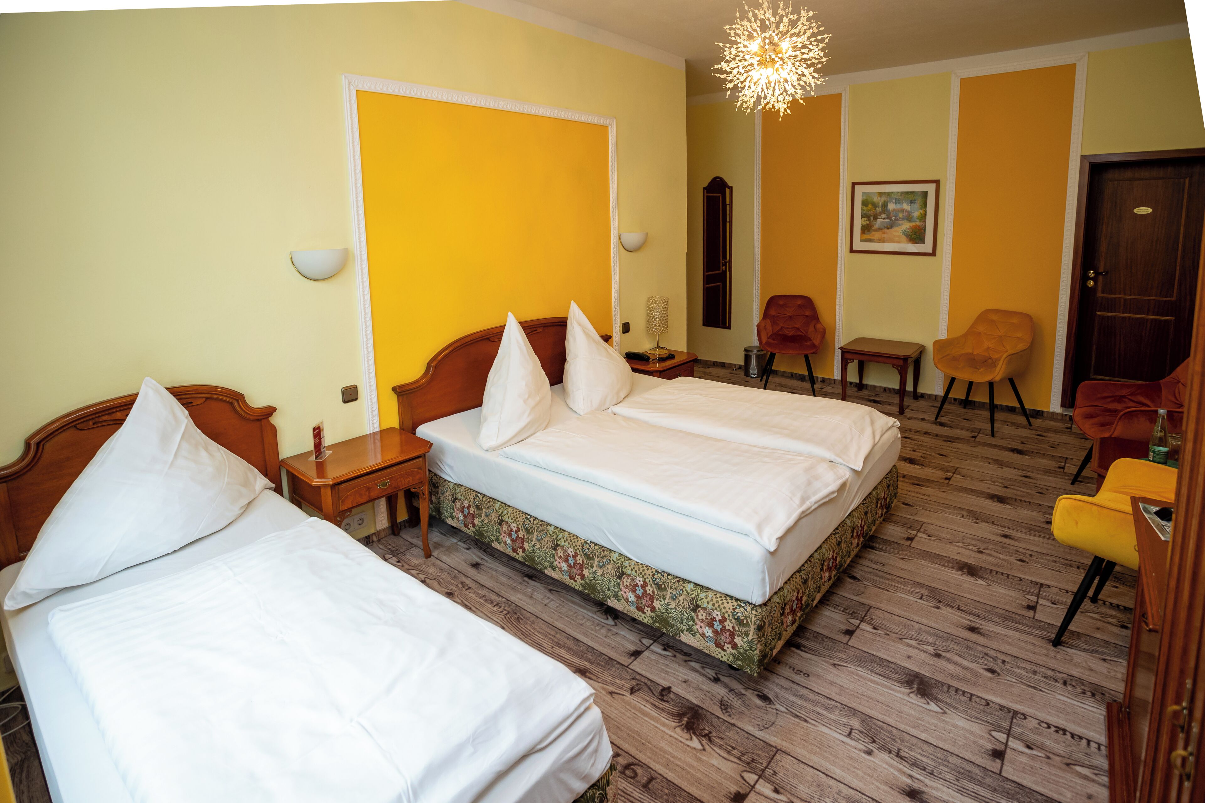 A hotel room with two beds, wooden flooring, a wooden headboard, a nightstand, a chair, a small table, a mirror, and a door.