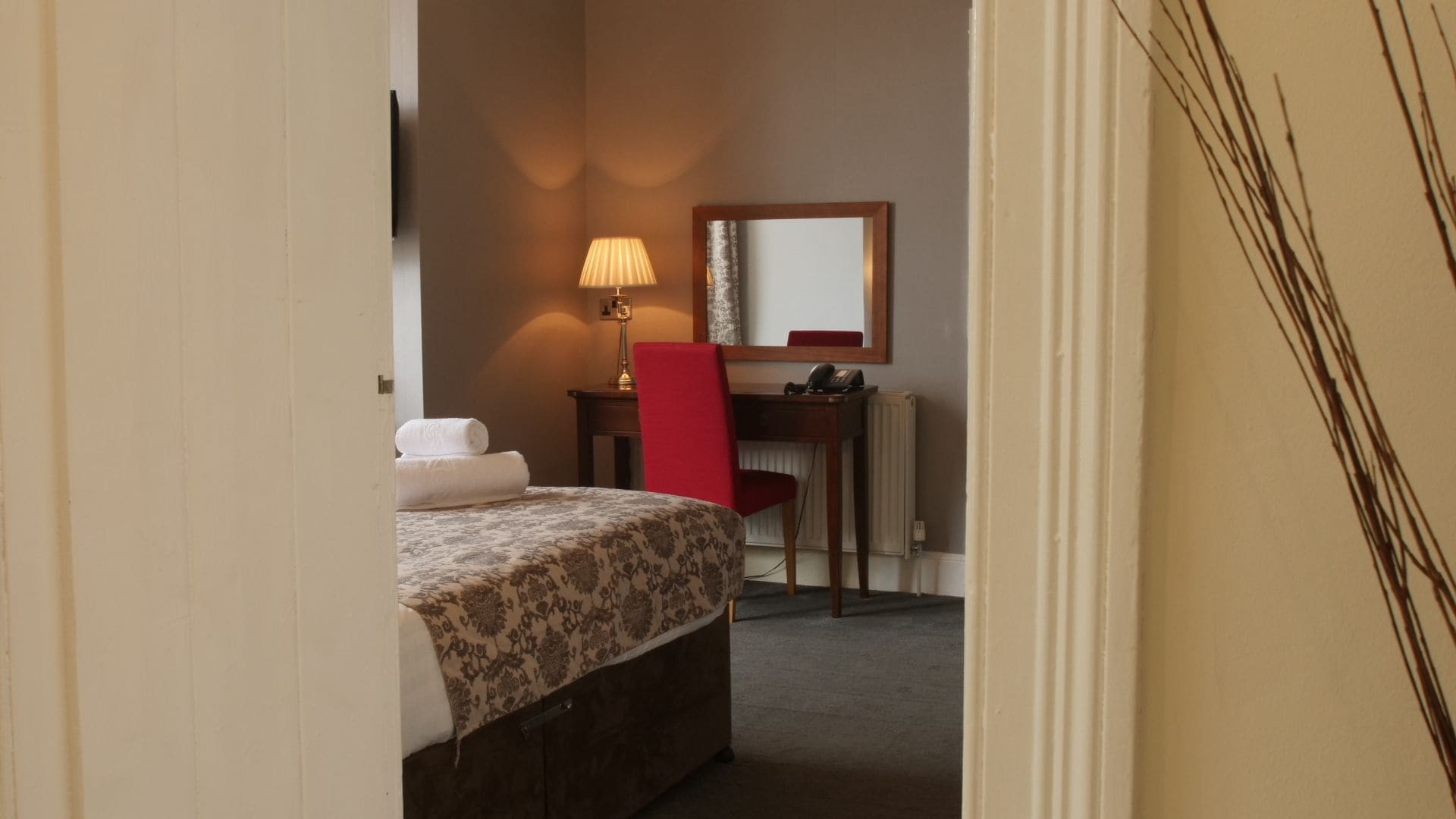 A hotel room with a bed, a desk, a chair, a lamp, and a radiator.