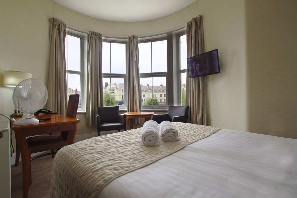 A hotel room with a bed, a desk with a lamp, a chair, a TV, and a view of buildings through large windows.
