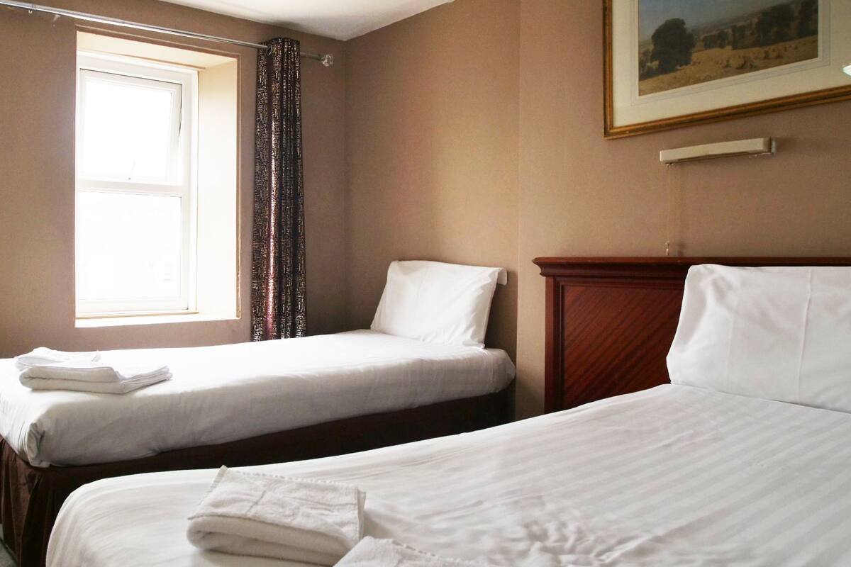 A hotel room with two single beds, a window with curtains, a painting on the wall, and a wooden headboard.