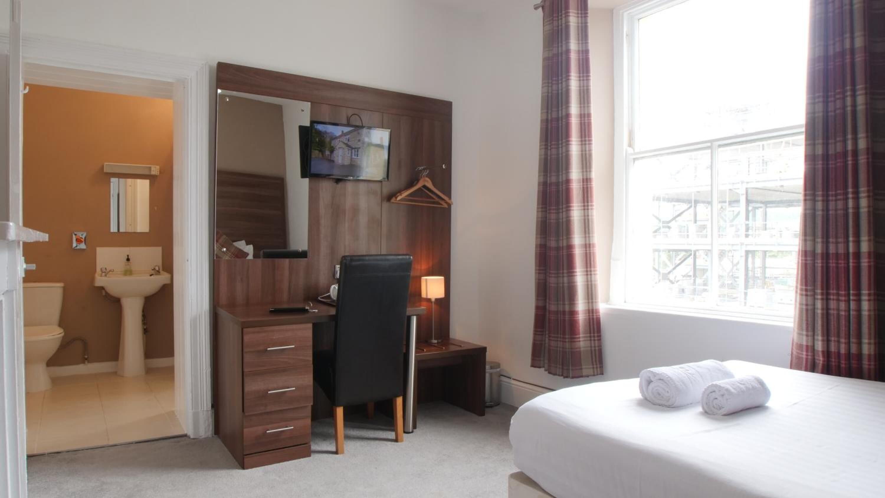 Une chambre d’hôtel comprenant un lit, un bureau avec une chaise, une télévision, une salle de bain avec un lavabo et une fenêtre avec des rideaux.