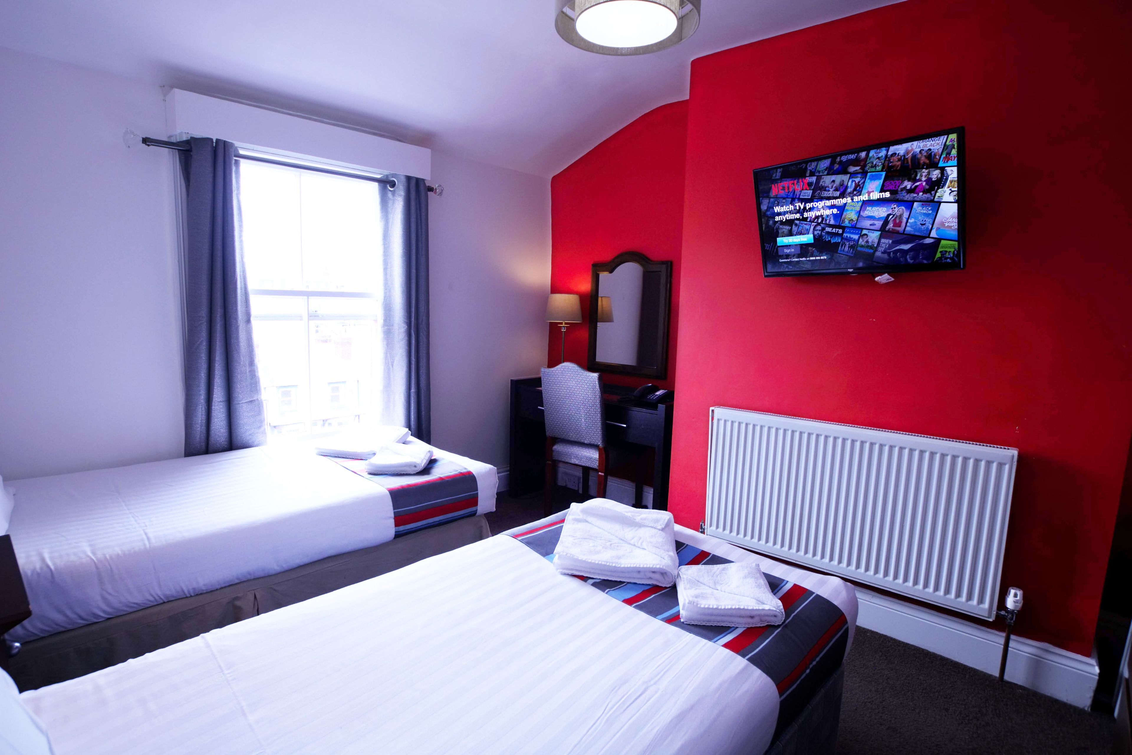 Une chambre d’hôtel avec deux lits, un téléviseur fixé au mur, un bureau et une fenêtre avec des rideaux.