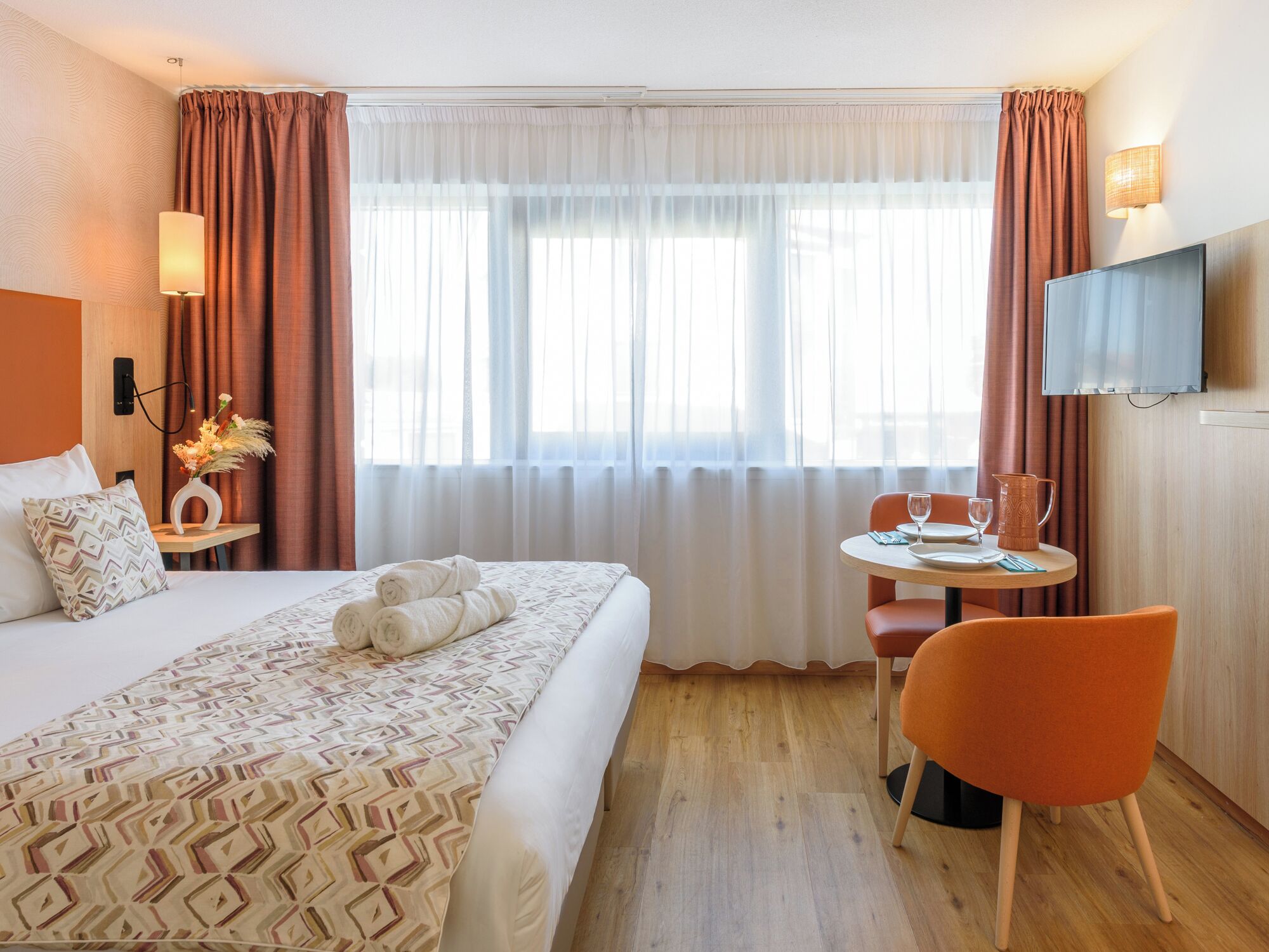 A hotel room with a bed, a small table with a chair, a TV, and a window with curtains.