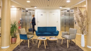A lobby with a blue sofa, two armchairs, a small table with magazines, a potted plant, and a fire extinguisher.