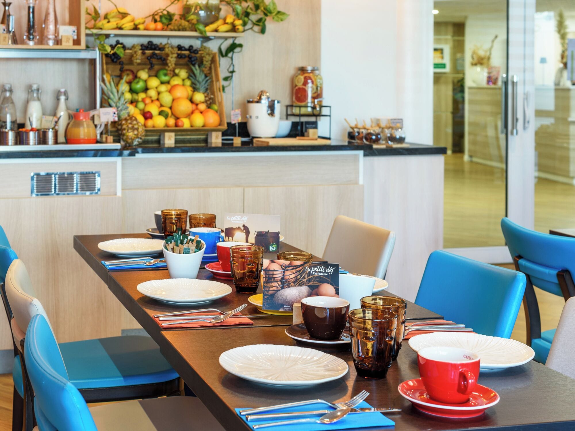 Un comedor con una mesa larga dispuesta para una comida, rodeada de sillas azules. Se ve una cocina con fruta y bebidas al fondo.